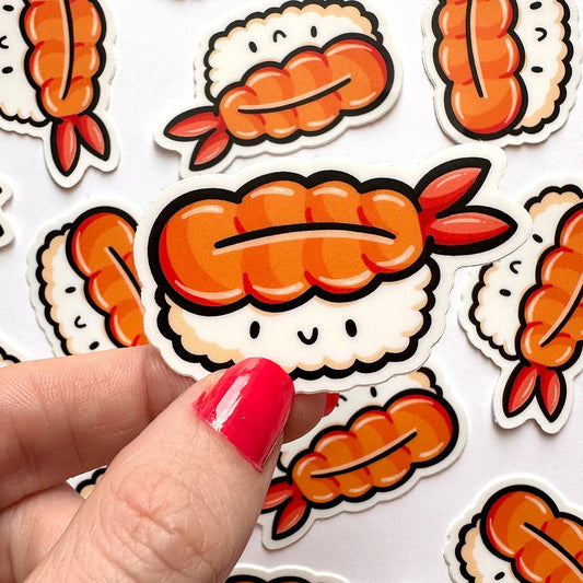 A close-up of a hand with bright pink nail polish holding a cute cartoon shrimp sushi sticker, with several matching sushi stickers scattered on a white background.