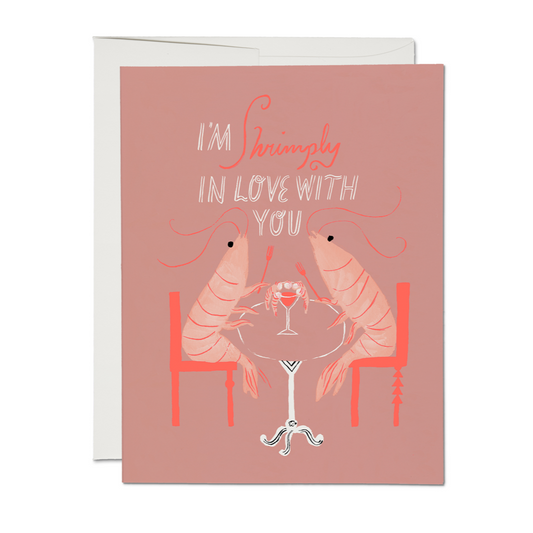 Pink greeting card with illustration of shrimps at dinner holding forks over a shared shrimp cocktail. Text reads "I'm shrimply in love with you" in fanciful writing.