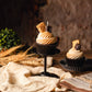 Realistic Smores Candle with chocolate cupcake base, whipped marshmallow topping and topped with a piece of chocolate and piece of graham cracker. More candle is on a cupcake base on pedestal. Table is autumnal. There is a second Smores candle in the distance.