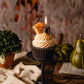 Realistic Smores Candle with chocolate cupcake base, whipped marshmallow topping and topped with a piece of chocolate and piece of graham cracker. SMore candle is on a cupcake base (lit) on pedestal. Table is autumnal. There is a second Smores candle in the distance.