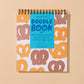 A spiral-bound Doodle Book with a bright blue cover and a white cover patterned with pretzels in orange, brown, and beige, displayed on a solid beige background.