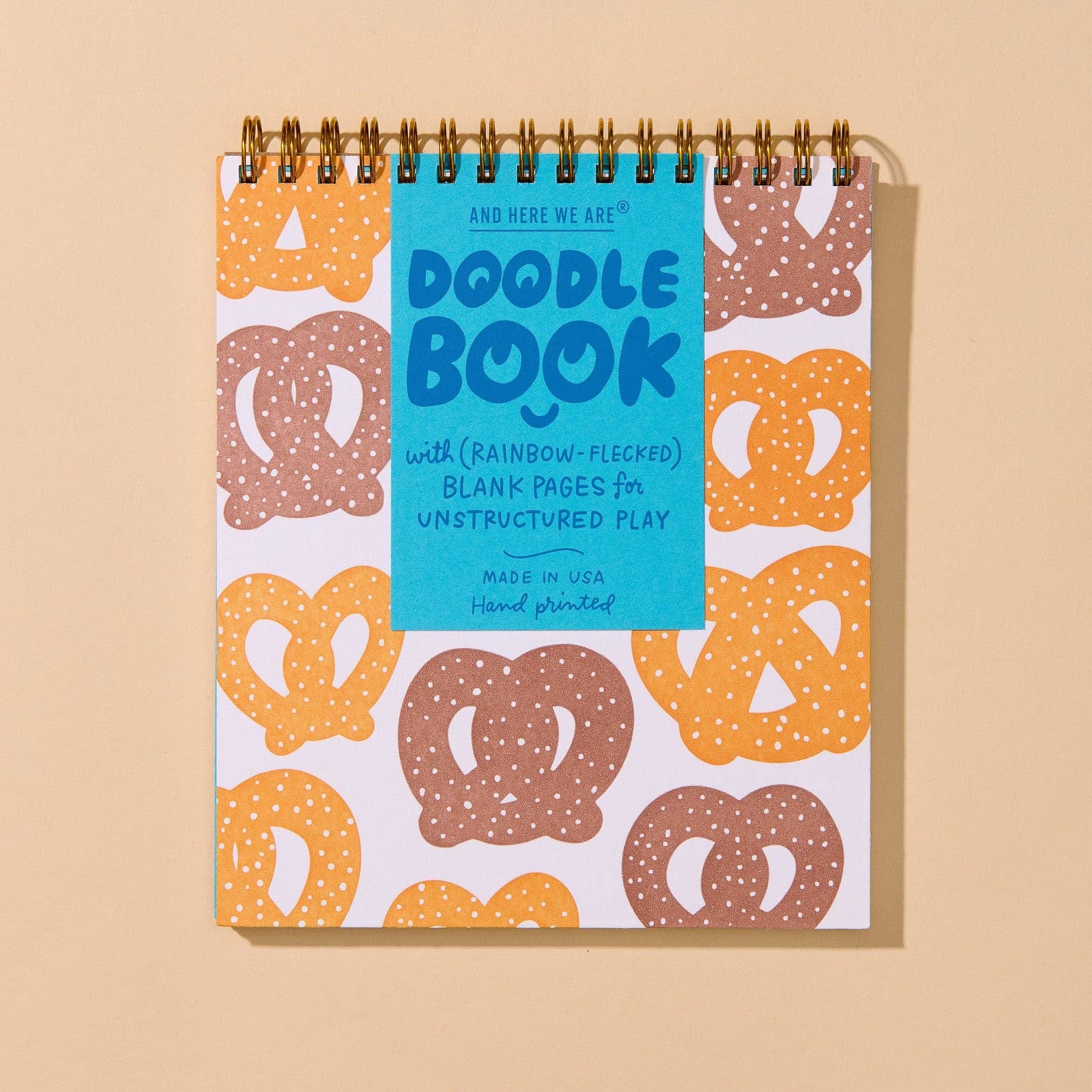A spiral-bound Doodle Book with a bright blue cover and a white cover patterned with pretzels in orange, brown, and beige, displayed on a solid beige background.