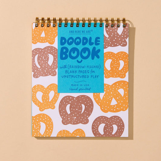 A spiral-bound Doodle Book with a bright blue cover and a white cover patterned with pretzels in orange, brown, and beige, displayed on a solid beige background.