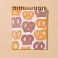 A spiral-bound notebook with a white cover decorated with a repeating pattern of pretzels in orange and brown, set against a solid beige background.