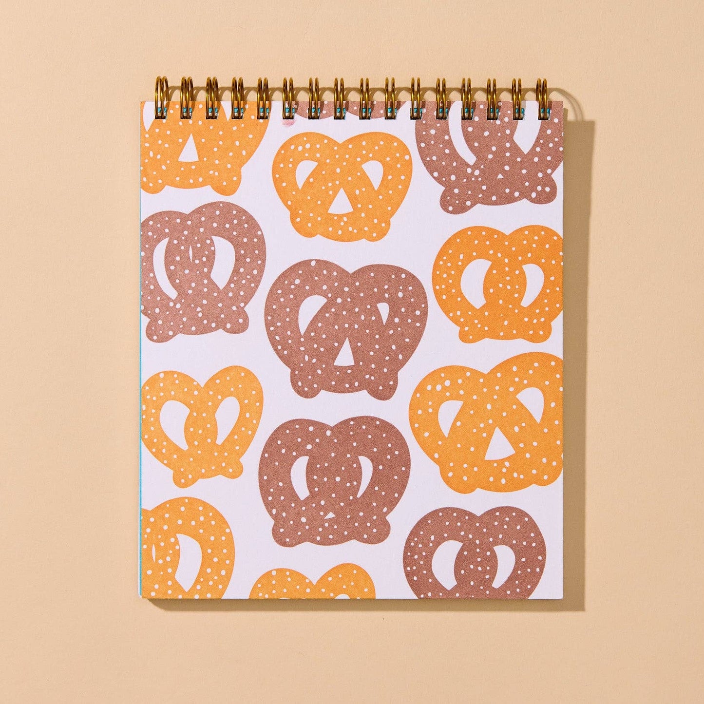A spiral-bound notebook with a white cover decorated with a repeating pattern of pretzels in orange and brown, set against a solid beige background.