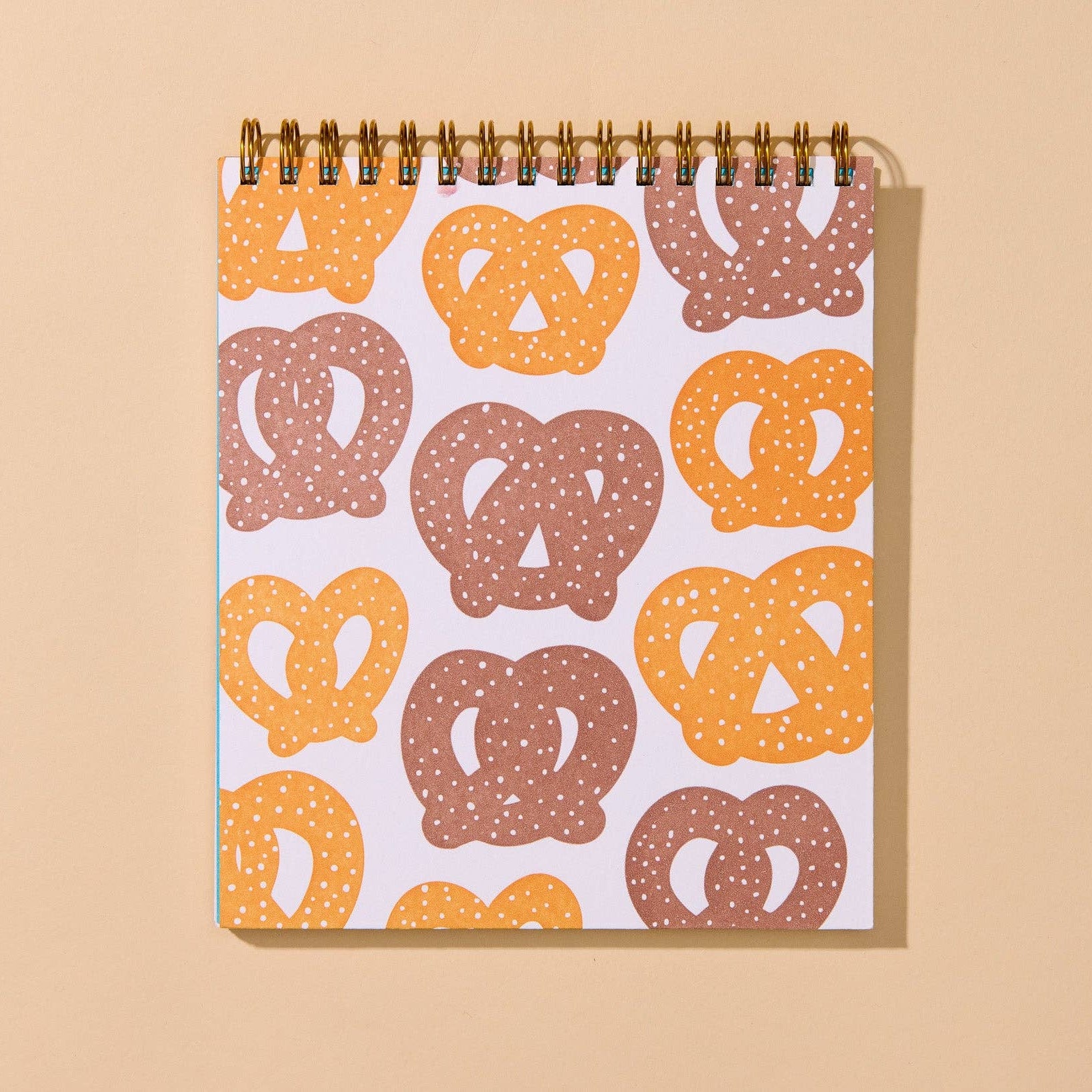A spiral-bound notebook with a white cover decorated with a repeating pattern of pretzels in orange and brown, set against a solid beige background.