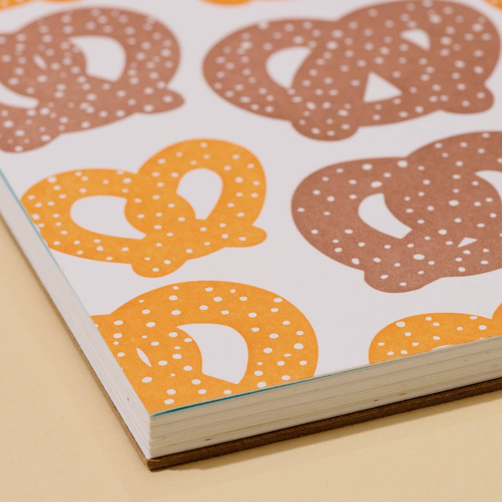 A close-up of a printed paper item, likely a notebook cover or cardstock sheet, featuring a repeating pattern of pretzels in brown and orange with white speckles on a white background.