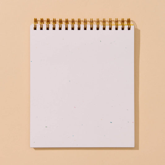 A blank white spiral-bound notepad with a gold wire binding, standing upright against a solid warm beige background.