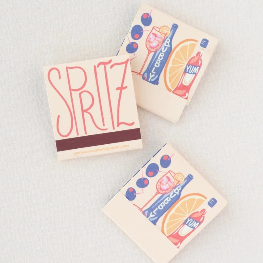 Spritz matchbook open to reaveal that the matches are in the shape of Campari, orange wedge, bubbles, spritz in a glass and olives on a pick.