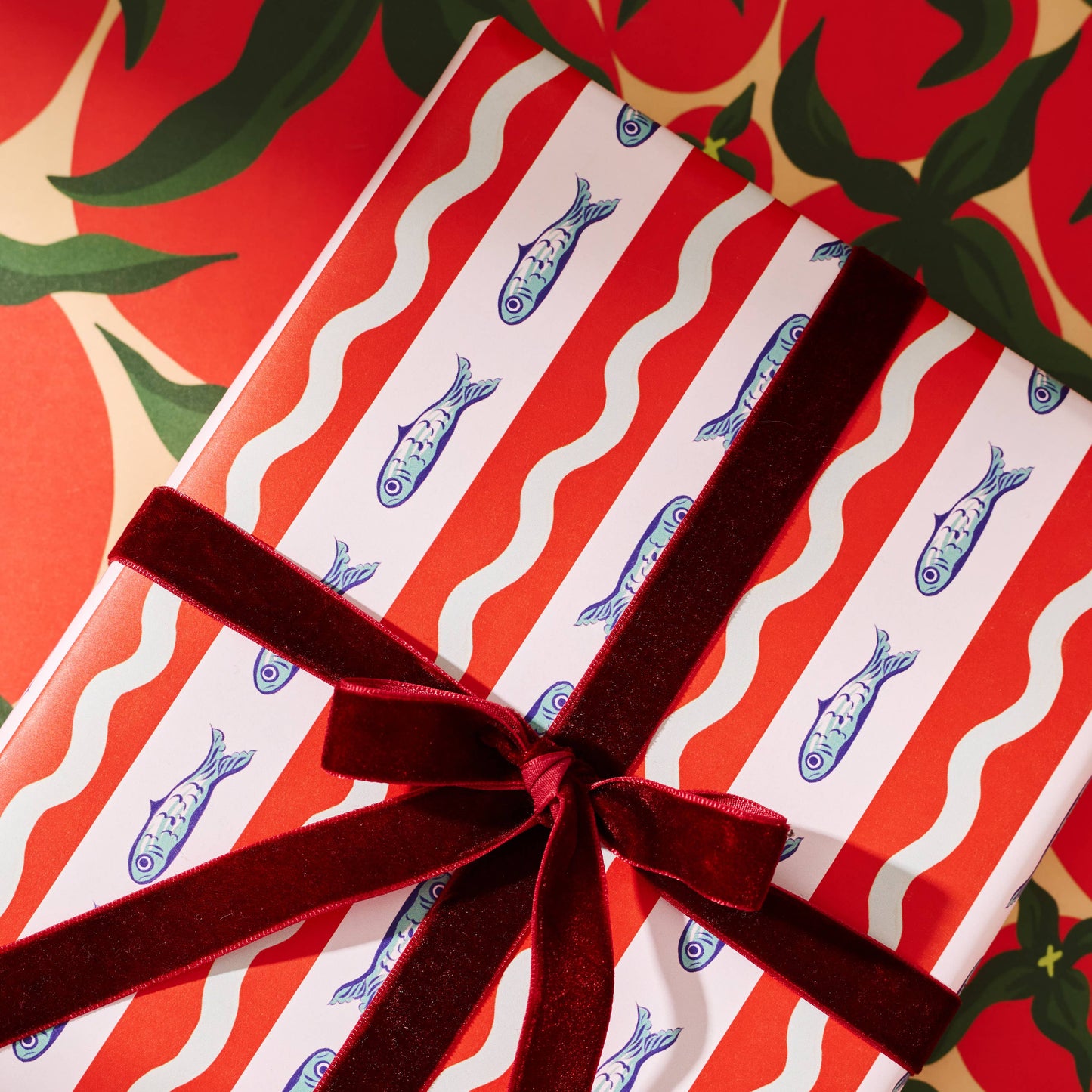 Gift wrapped in red and white striped paper with blue fish patterns, tied with a red ribbon, against a red background with green leaves.