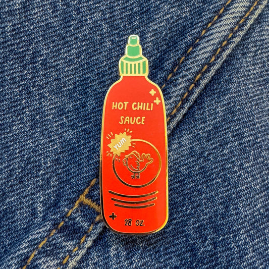 A chili sauce bottle-shaped enamel pin with a red body, green cap, and yellow sunburst graphic, pinned onto blue denim fabric.
