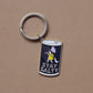 A silver metal keychain with a cylindrical charm featuring a navy blue background, a cartoon cat in a yellow raincoat, and the words 'STAY SALTY' in white, shown on a plain brown background.