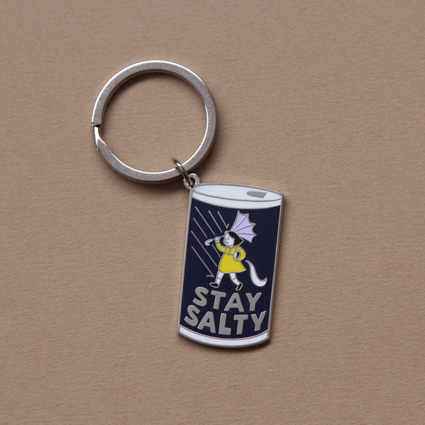 A silver metal keychain with a cylindrical charm featuring a navy blue background, a cartoon cat in a yellow raincoat, and the words 'STAY SALTY' in white, shown on a plain brown background.