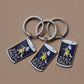 Three enamel keychains with silver metal rings and cylindrical charms featuring a navy blue background, a white cat in a yellow dress, and the text 'STAY SALTY' on a plain tan surface.