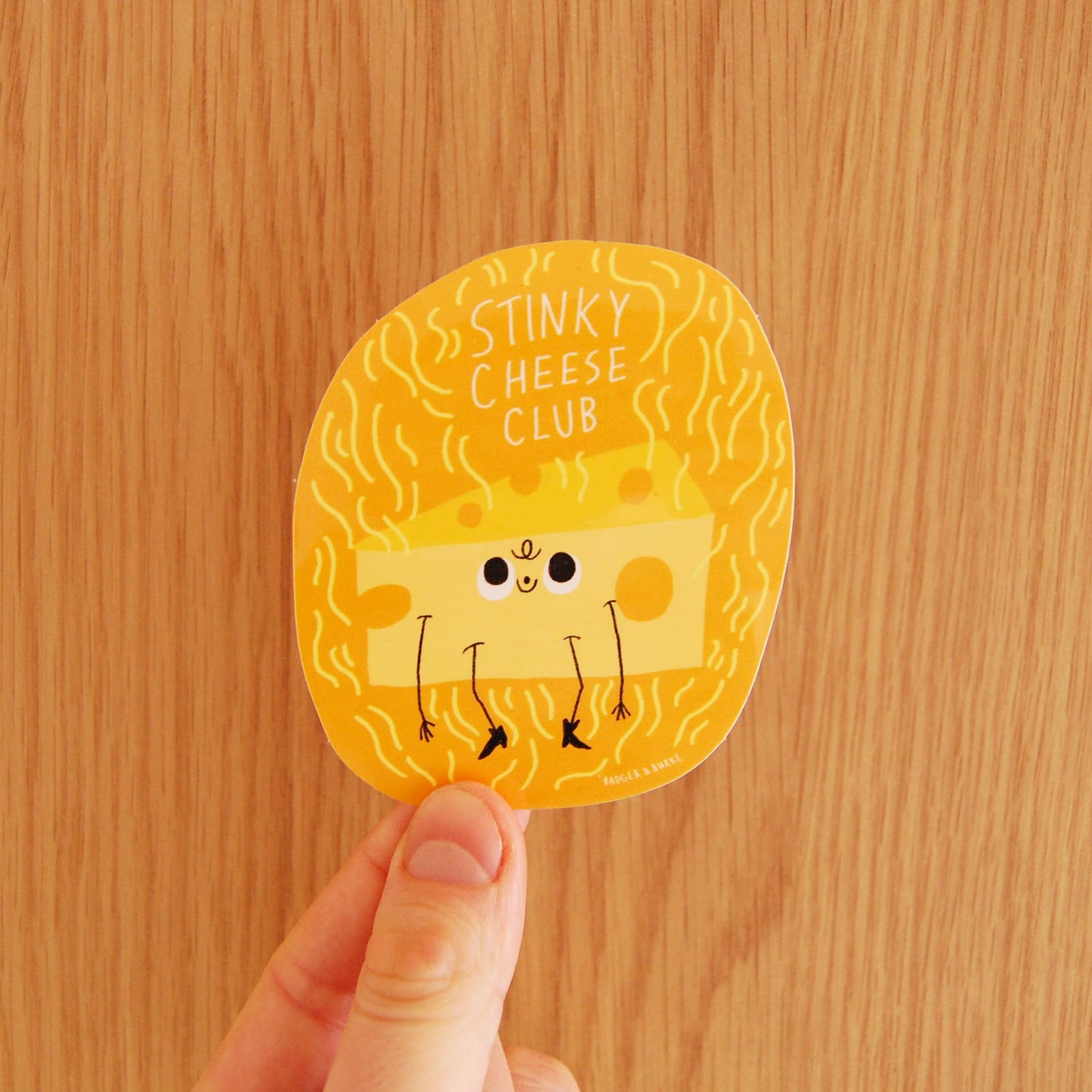 Round sticker with cartoon cheese character and 'Stinky Cheese Club' text held against a wooden background