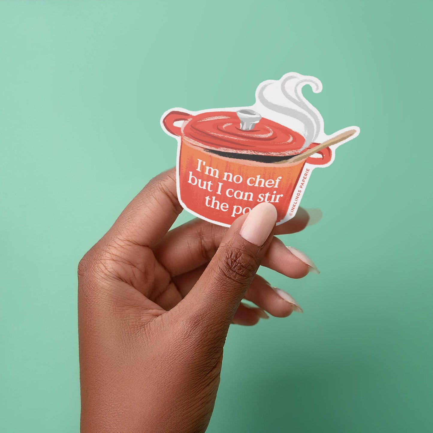 Hand holding a sticker of a pot with text against a green background