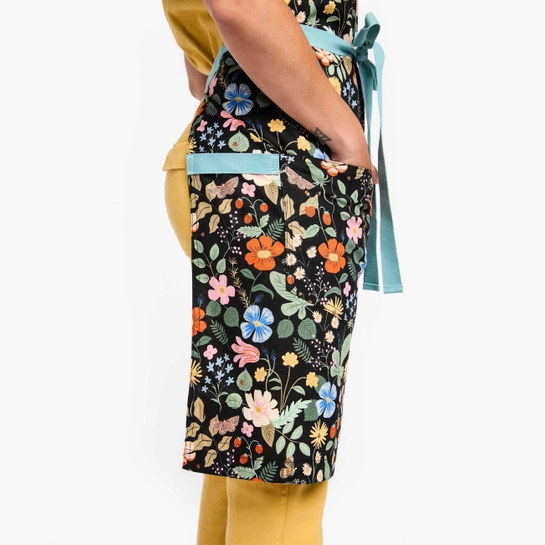Side view of apron with colorful design pattern of bright florals and foliage and light blue neck and waist straps 
