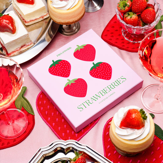 Strawberry-themed table setting with desserts and drinks on a pink background. Box of linen napkins on display.