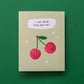 A square greeting card with a light beige background, featuring an illustration of two smiling red cherries with green stems and a speech bubble that reads 'I LOVE BEING STUCK WITH YOU!'. The card is photographed flat against a solid dark green background.