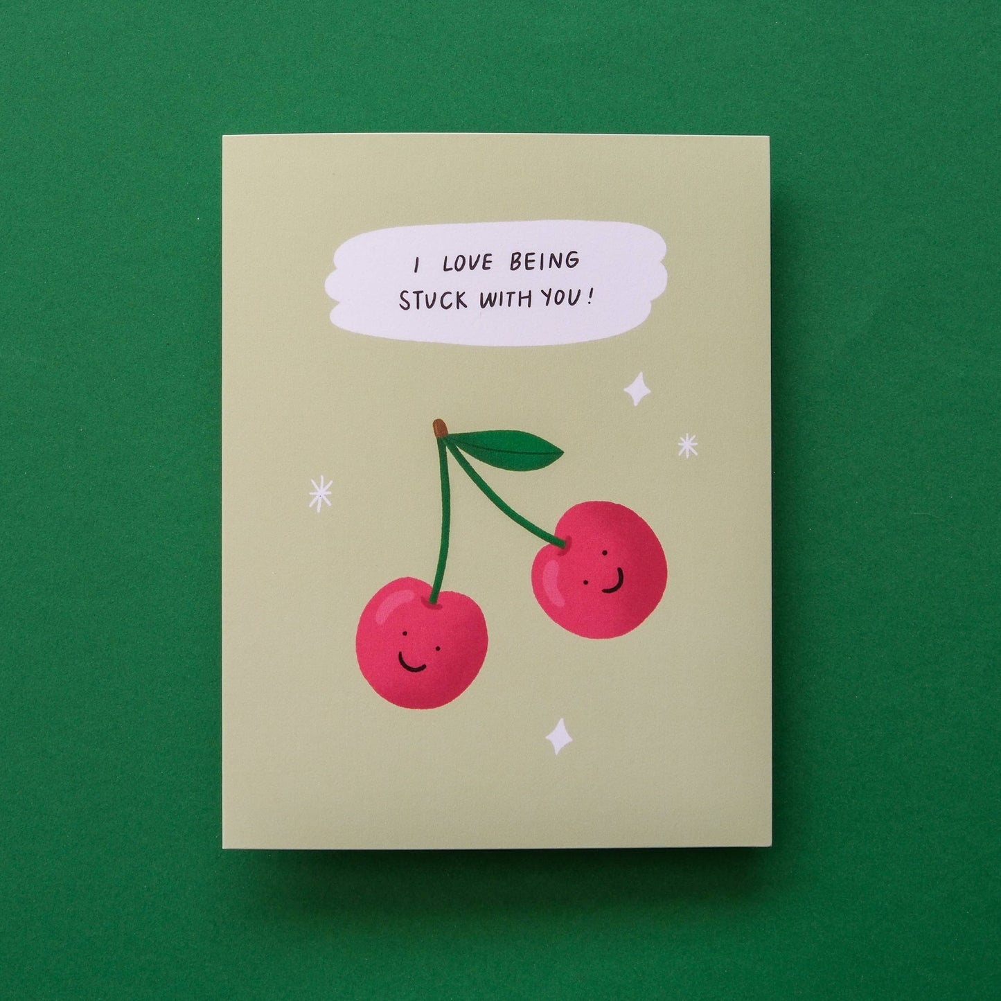 A square greeting card with a light beige background, featuring an illustration of two smiling red cherries with green stems and a speech bubble that reads 'I LOVE BEING STUCK WITH YOU!'. The card is photographed flat against a solid dark green background.