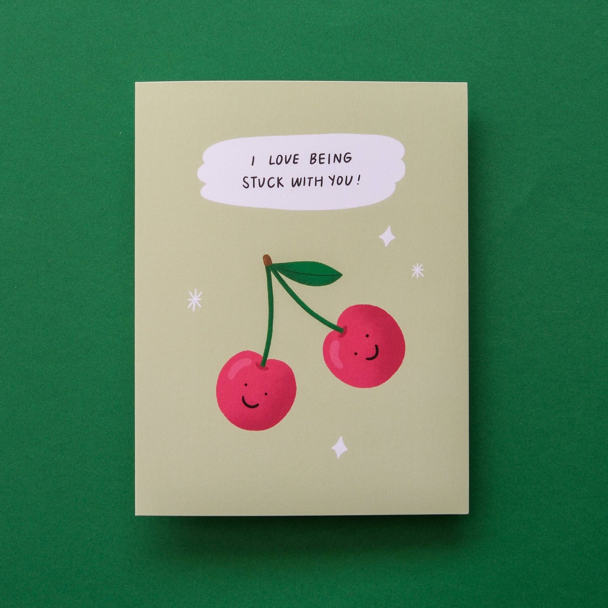 A square greeting card with a light beige background, featuring an illustration of two smiling red cherries with green stems and a speech bubble that reads 'I LOVE BEING STUCK WITH YOU!'. The card is photographed flat against a solid dark green background.