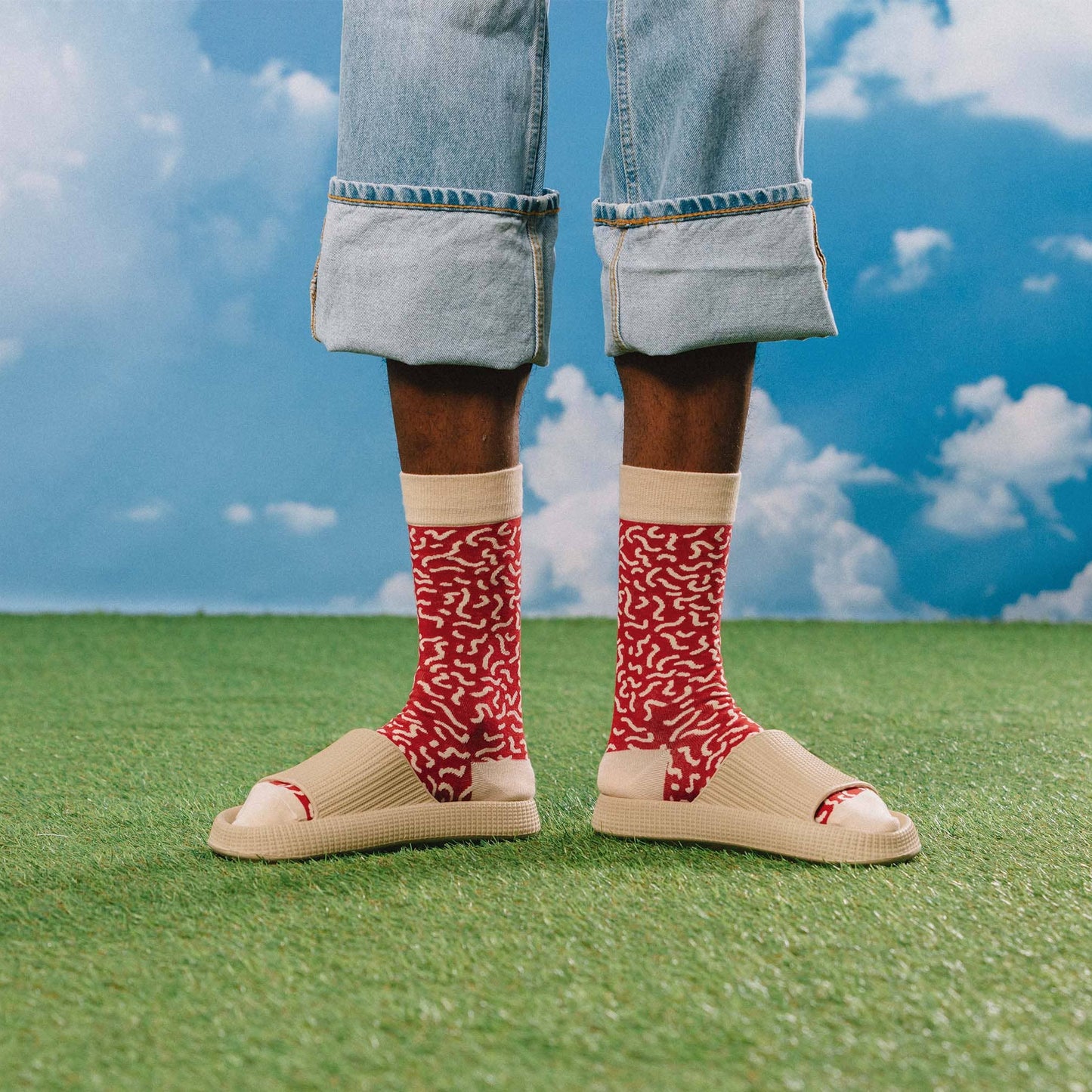 Person wearing red patterned socks and beige sandals on grass with a blue sky background