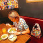 Silver bag featuring several popular Thai food dishes and text written in Thai. A Mui Thai boxer is on the bottom right.  Girl eating Thai food at a restaurant with the bag next to her in the booth.
