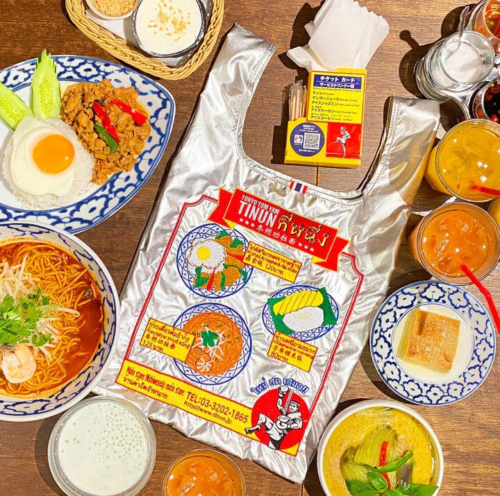 Silver bag featuring several popular Thai food dishes and text written in Thai. A Mui Thai boxer is on the bottom right.  The dishes that are embroidered on the bag are on the table for showcasing.