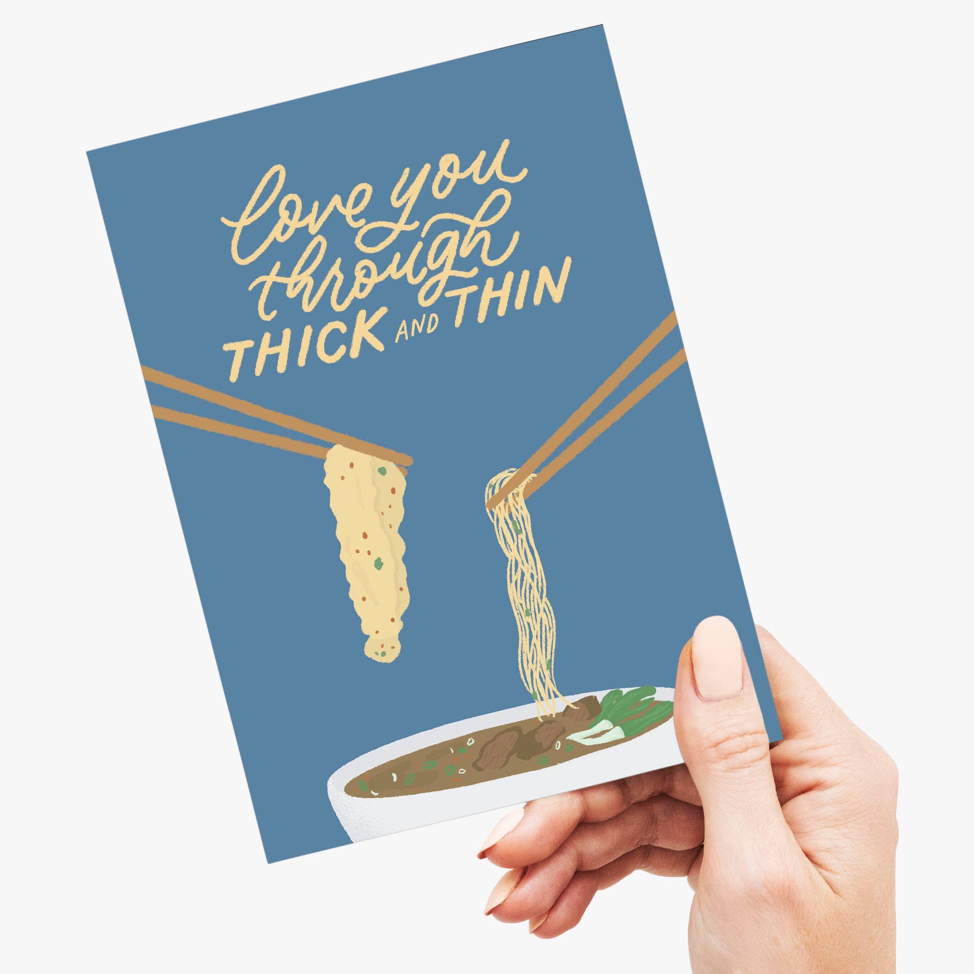 Card with noodles and text 'love you through thick and thin' held by a hand on a white background