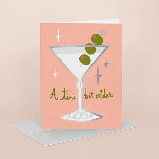 A pink greeting card with a martini glass illustration, green olives, and the phrase 'A tini bit older' in gold script, shown standing upright with a white envelope behind it on a pale pink background.