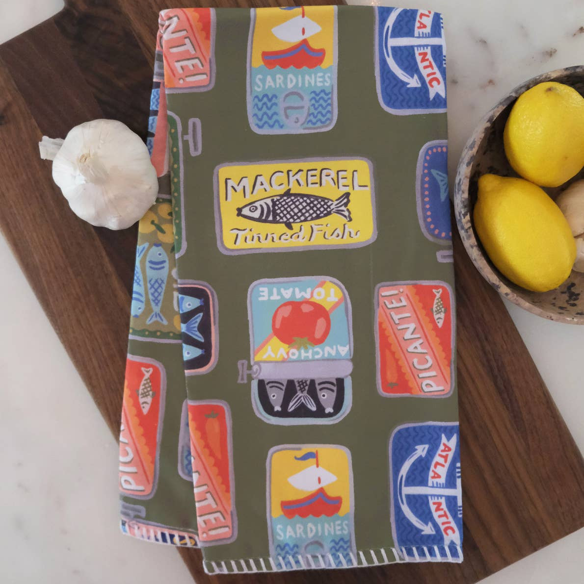 Patterned kitchen towel with vintage tinned fish label design on a wooden cutting board.