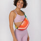 Fanny pack that looks like a tomato slice with seeds. Orange, red and yellow bag with green straps. Woman wearing the pack as a cross body sling.
