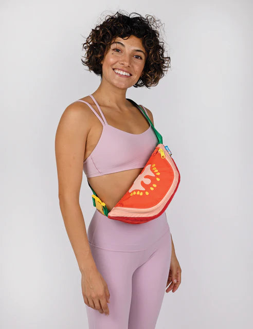 Fanny pack that looks like a tomato slice with seeds. Orange, red and yellow bag with green straps. Woman wearing the pack as a cross body sling.