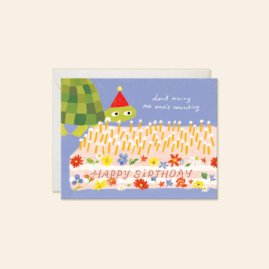 birthday card with light blue background and illustration of a turtle wearing a party hat and a large sheet cake decorated with flowers and text that says "happy birthday" and has lots of candles on top. Card reads "don't worry no one's counting" on the outside 