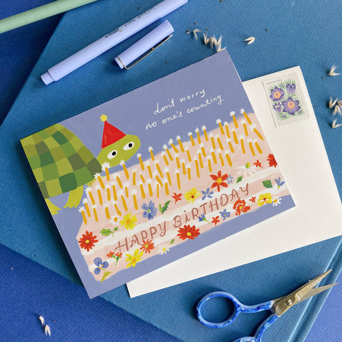 birthday card with light blue background and illustration of a turtle wearing a party hat and a large sheet cake decorated with flowers and text that says "happy birthday" and has lots of candles on top. Card reads "don't worry no one's counting" on the outside 