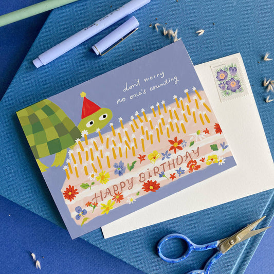 birthday card with light blue background and illustration of a turtle wearing a party hat and a large sheet cake decorated with flowers and text that says "happy birthday" and has lots of candles on top. Card reads "don't worry no one's counting" on the outside 