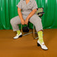 Person sitting on a chair wearing a checkered outfit with green socks against a green curtain background
