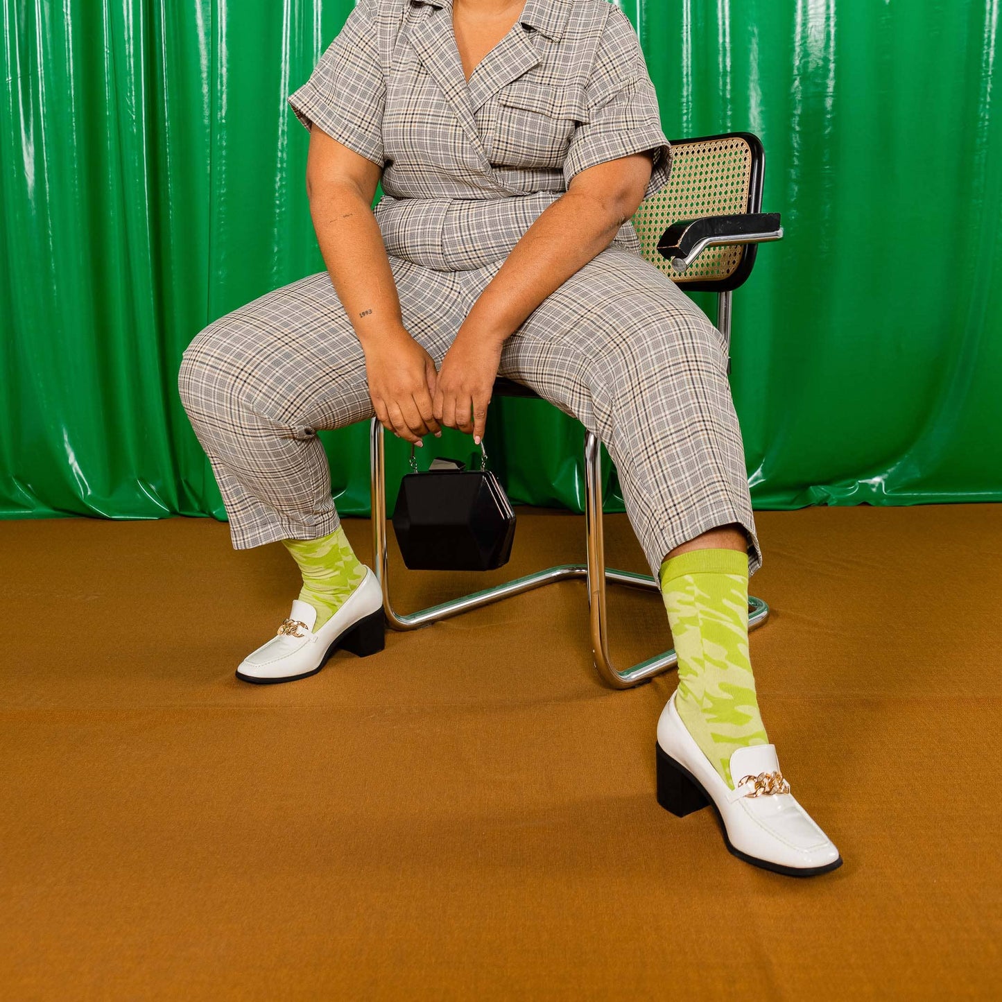 Person sitting on a chair wearing a checkered outfit with green socks against a green curtain background
