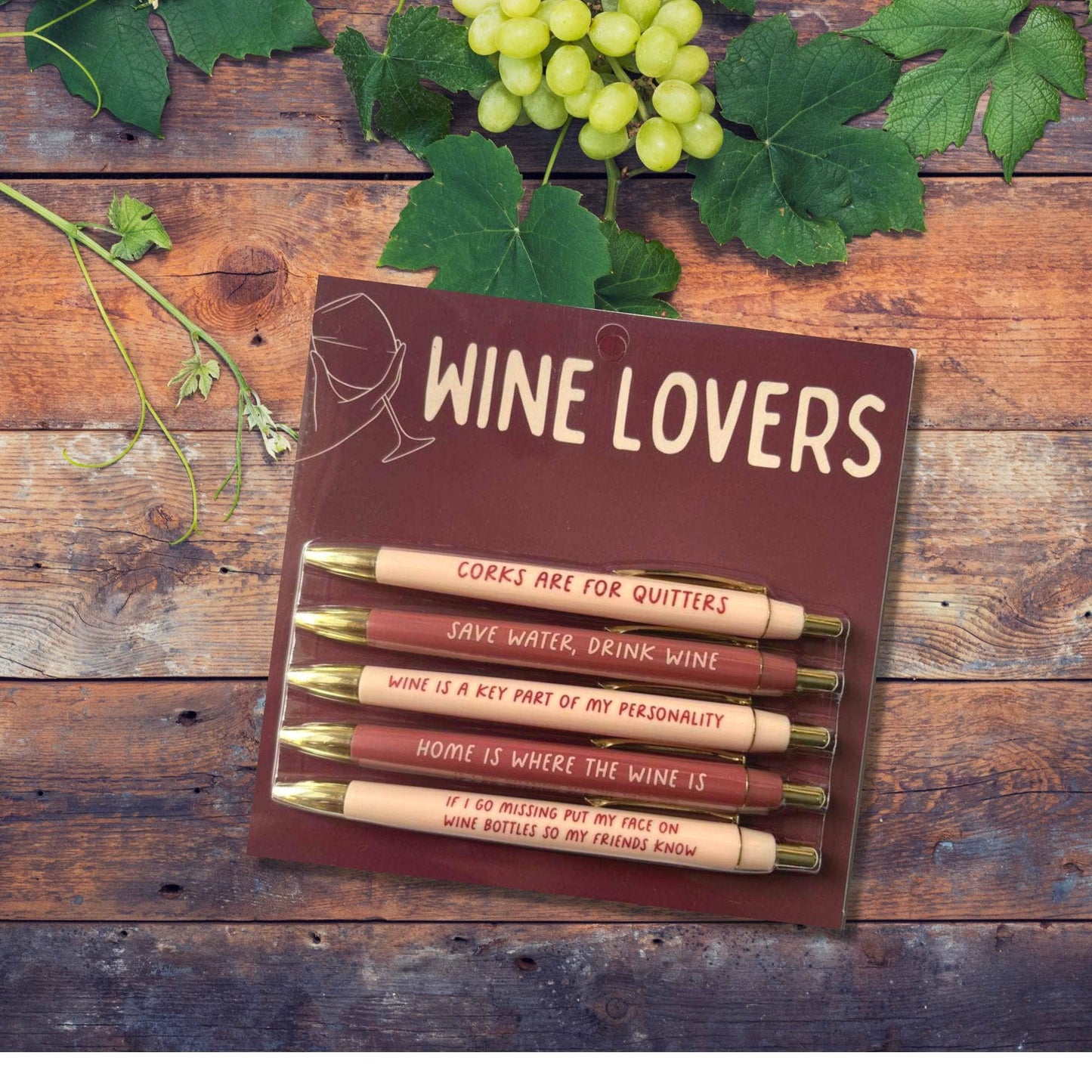 Pack of wine-themed pens with humorous sayings on a wooden surface with grapes and leaves.