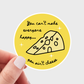 Sticker with cheese illustration and text held by a hand on a light gray background