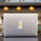 A silver laptop on a dark tabletop with a blurred indoor background and warm hanging lights. The laptop lid has a small sticker of a butter stick with the text 'You Butter Twerk'.