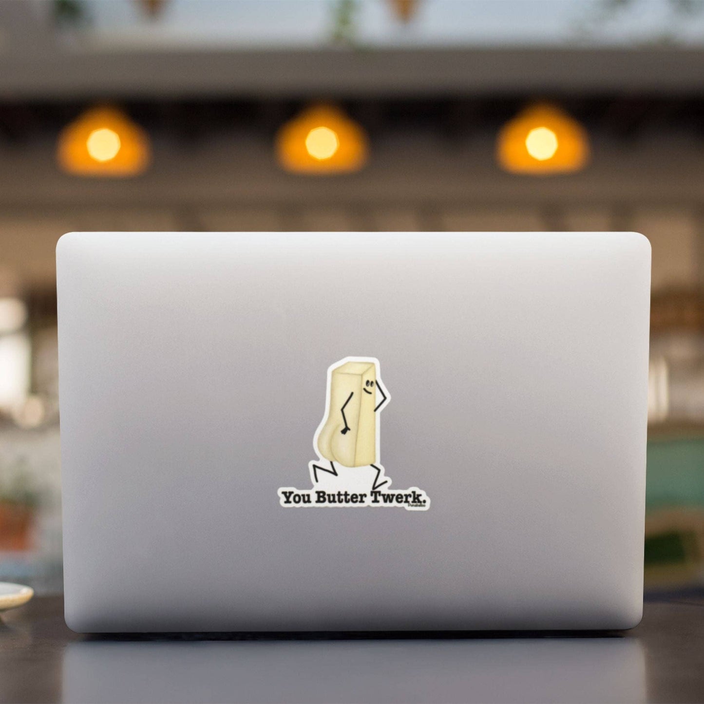 A silver laptop on a dark tabletop with a blurred indoor background and warm hanging lights. The laptop lid has a small sticker of a butter stick with the text 'You Butter Twerk'.