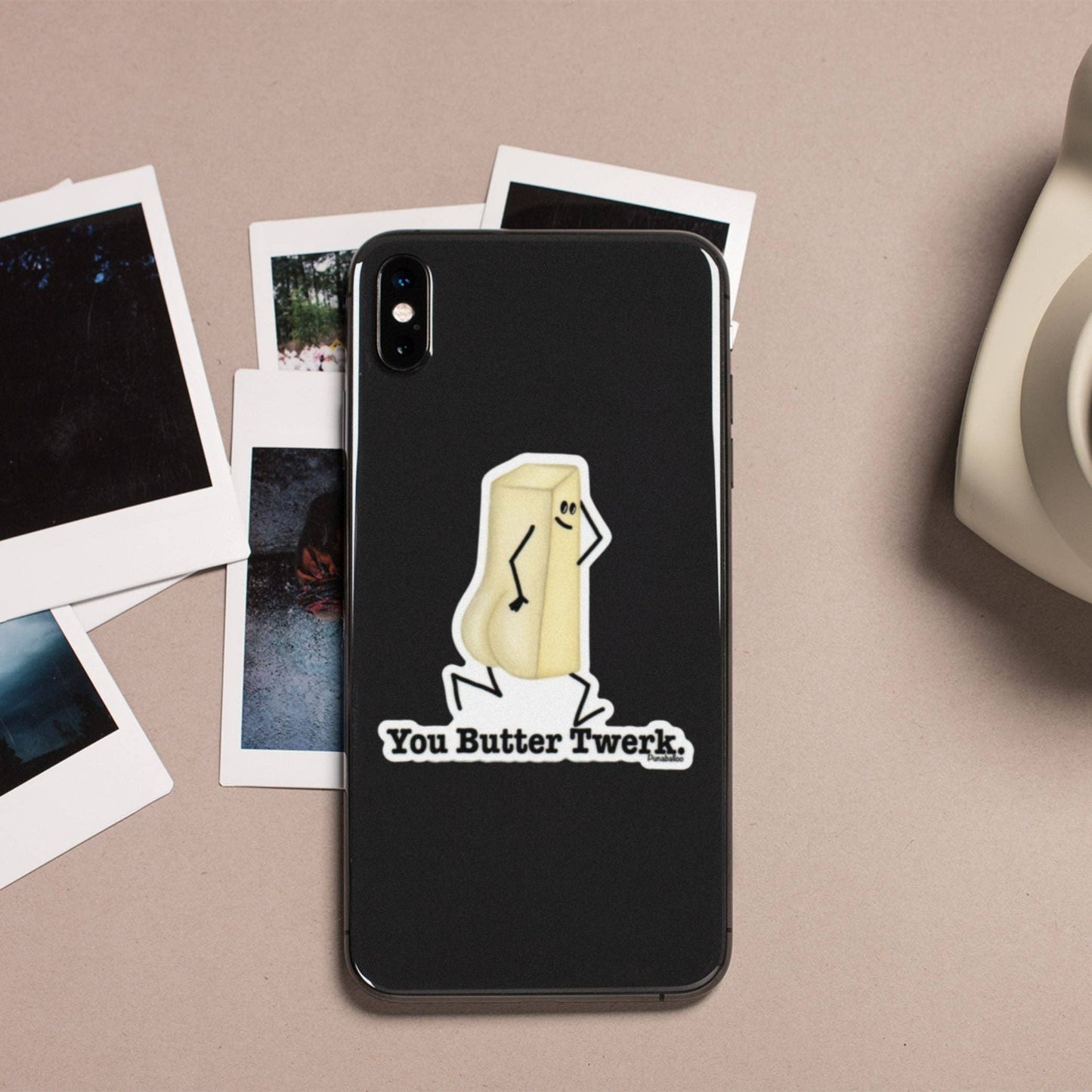 A black phone case featuring a cartoon butter character and the text 'You Butter Twerk.' displayed on a beige surface with scattered Polaroid-style photos and a partial white mug nearby.