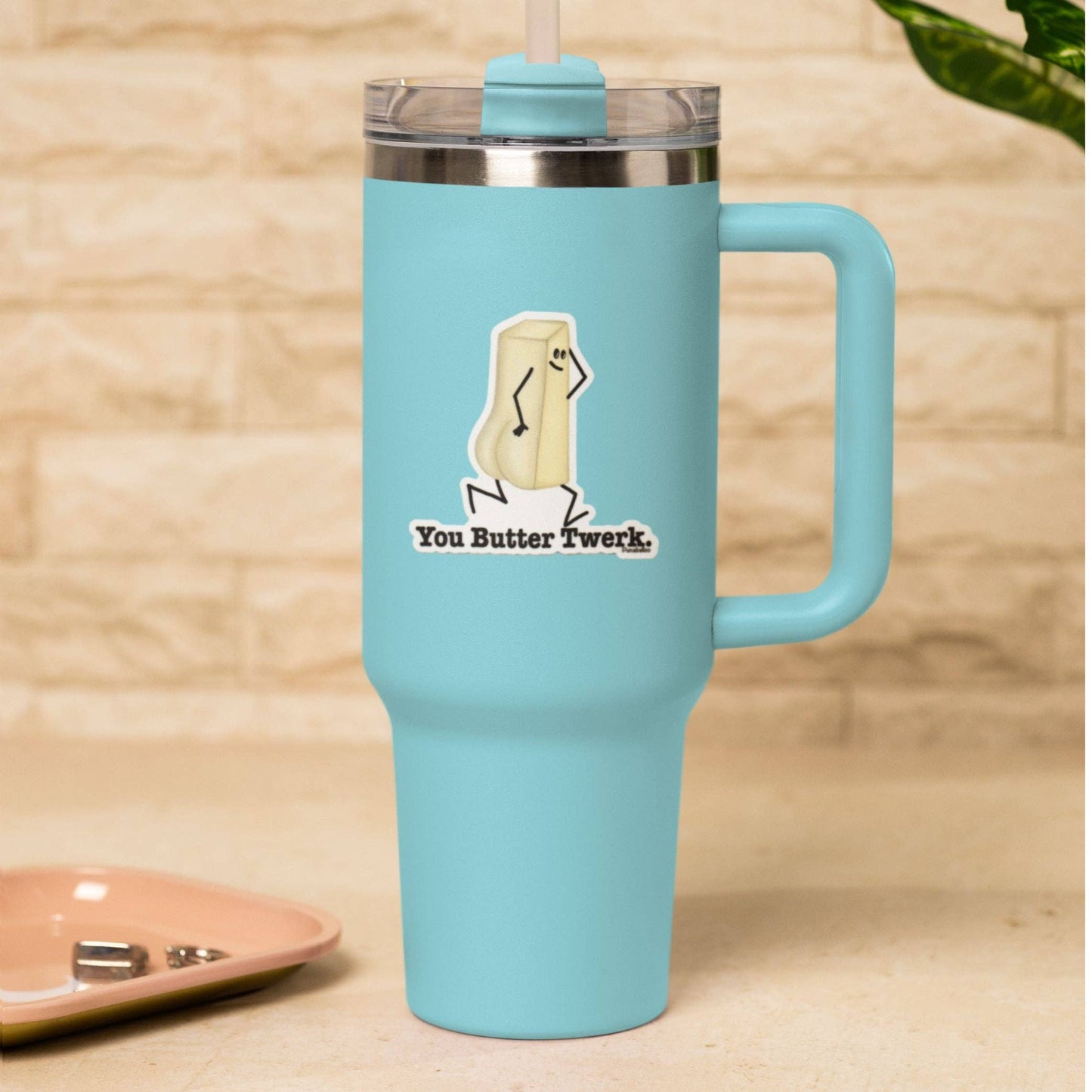 A light blue insulated travel tumbler with a handle, clear lid, and straw, featuring a cartoon butter stick graphic and the text 'You Butter Twerk.' on the front, set against a warm beige brick wall background.