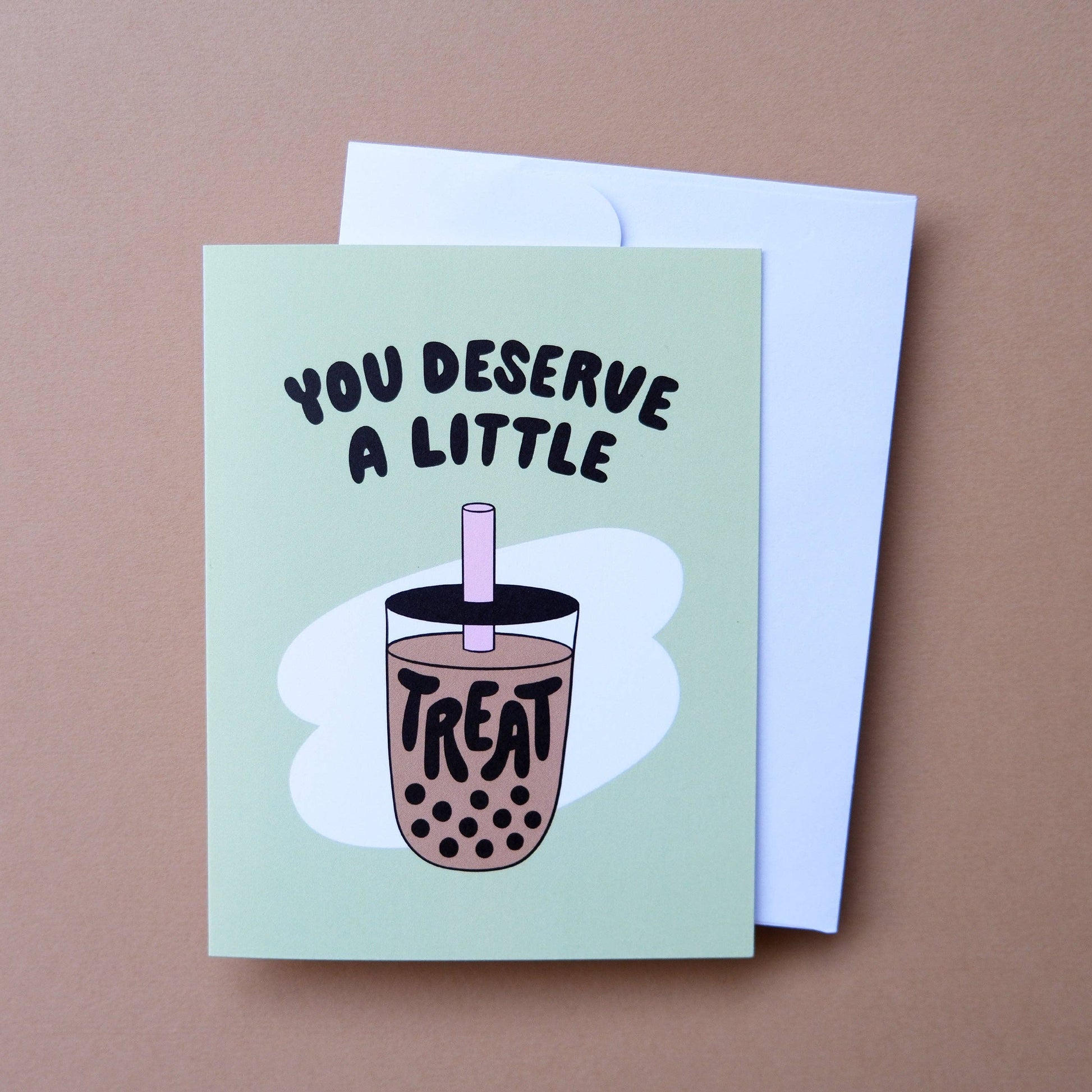 A square greeting card with a light mint green background, featuring a hand-drawn illustration of a boba tea cup with the text 'YOU DESERVE A LITTLE TREAT'. A white envelope is placed behind the card on a solid brown surface.