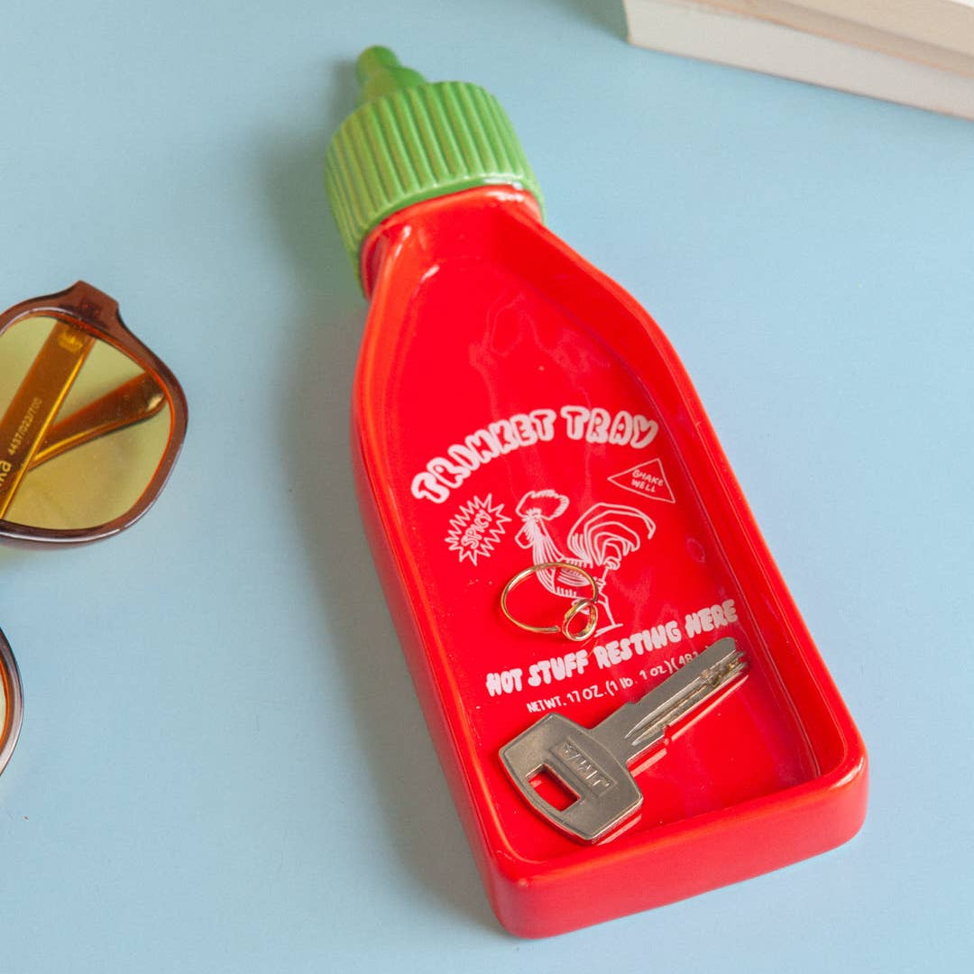 Red sriracha bottle-shaped trinket tray with a green cap on a light blue background, holding a ring and a key