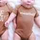 Two babies wearing brown onesies with text, sitting on a white surface.