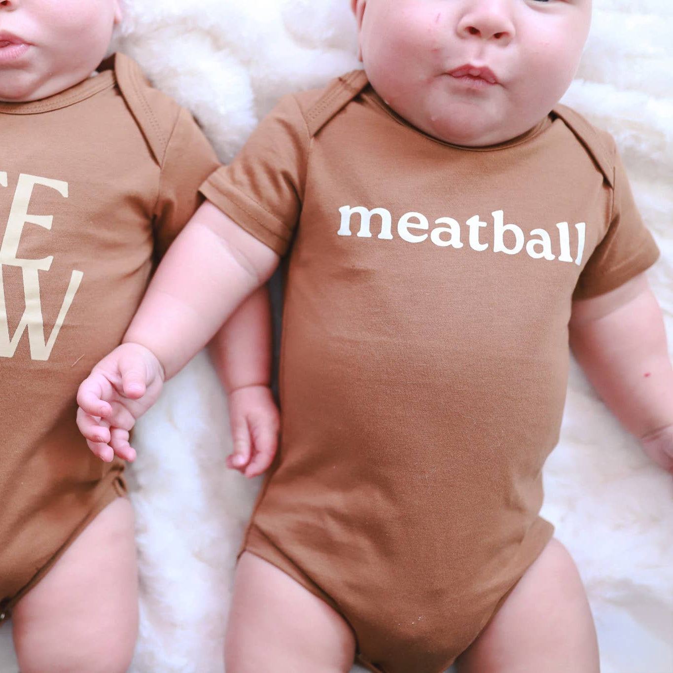 Two babies wearing brown onesies with text, sitting on a white surface.