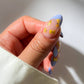 Hand with blue nail polish featuring yellow pasta designs on a neutral background