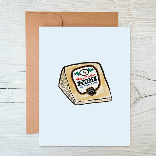 Greeting card with a cheese illustration on a wooden surface. Text on card reads "Here's another cheesy birthday card" 